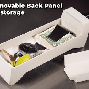 May include: A white plastic storage box with a removable back panel. The box is open and contains a money clip with cash, a key with a key fob, and a credit card. The text "STAY TIGHT" is printed on the side of the box.