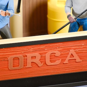 May include: A detailed model of the Orca fishing boat from the movie Jaws. The boat's side is a textured orange with the name "ORCA" in large letters. Figures of characters are visible, one holding a rifle and another with a pole.