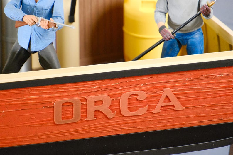May include: A detailed model of the Orca fishing boat from the movie Jaws. The boat's side is a textured orange with the name "ORCA" in large letters. Figures of characters are visible, one holding a rifle and another with a pole.