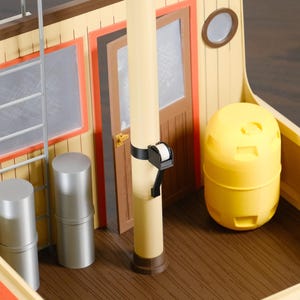 May include: A toy boat deck featuring a cream-colored wall with a brown door, a silver ladder, and a central mast. Two silver cylindrical canisters stand near the ladder, while a bright yellow barrel sits on the brown wood-grain floor.