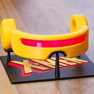 May include: A yellow visor with a horizontal red lens, mounted on a black display stand. The base of the stand features the text X-MEN in raised yellow letters with red shadowing, supported by two black vertical pillars holding the visor.