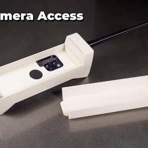 May include: A white plastic case with a black antenna, designed to protect a camera lens. The case is open, revealing the lens and a black circle in the center. The case is labeled "Camera Access".