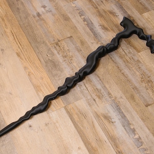 May include: A matte black walking cane featuring a distinctively wavy, organic silhouette throughout its entire length. The handle is shaped with a curved, ergonomic grip that transitions into a textured, undulating shaft that tapers to a point.
