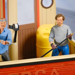 May include: Action figures of two men on a boat. One figure wears a blue shirt and holds a rifle. The other figure wears a gray shirt and holds a long pole. The boat is red with the word "ORCA" visible.