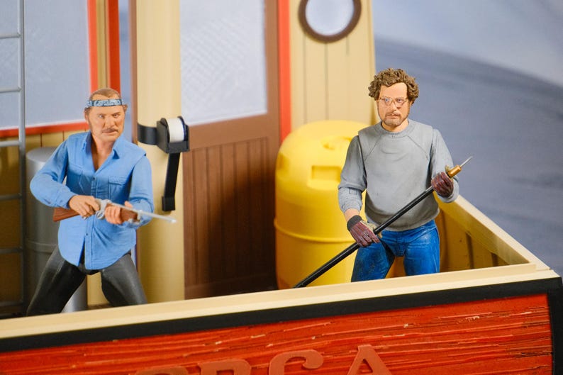 May include: Action figures of two men on a boat. One figure wears a blue shirt and holds a rifle. The other figure wears a gray shirt and holds a long pole. The boat is red with the word "ORCA" visible.