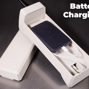 May include: A white plastic phone-shaped charging dock with a black wireless charging pad inside. The charging pad is connected to a white charging cable. The text "Battery Charging" is visible in the upper right corner of the image.