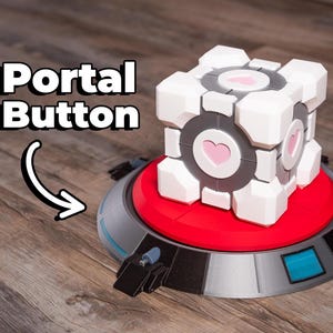 May include: A white and gray cube with pink heart accents, labeled "Portal Button," sits atop a red and gray circular base. The base has a blue and black design. The image is on a wooden surface.