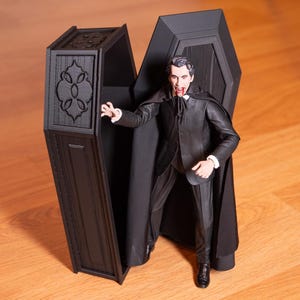 Peut inclure: Un cercueil en plastique noir avec un motif détaillé sur le côté, ouvert pour révéler une figurine d'action de vampire avec une cape noire et une tache rouge sur la bouche.