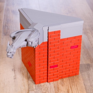 May include: A corner-shaped structure featuring a red brick wall pattern with a light gray top ledge. A light gray gargoyle figure extends from the corner of the ledge. The object has a triangular top surface and a vertical orientation.