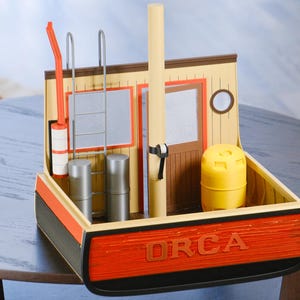 May include: A detailed model of a boat with the word "ORCA" in red lettering on the front. The model features a yellow barrel, a ladder, and a tall, light-colored pole. The boat's exterior is painted in shades of orange, black, and tan.