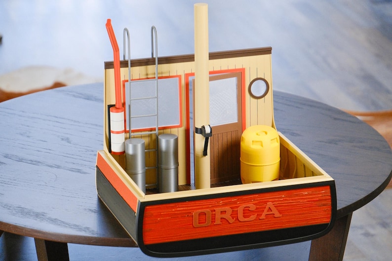 May include: A detailed model of a boat with the word "ORCA" in red lettering on the front. The model features a yellow barrel, a ladder, and a tall, light-colored pole. The boat's exterior is painted in shades of orange, black, and tan.