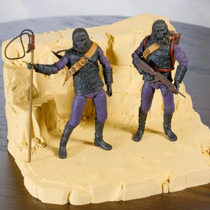 May include: Two ape-like action figures in purple and black attire, posed on a tan, textured base. One figure holds a staff with a loop, the other a rifle. The figures have detailed facial features and are positioned as if in action. The base is a light beige colour.