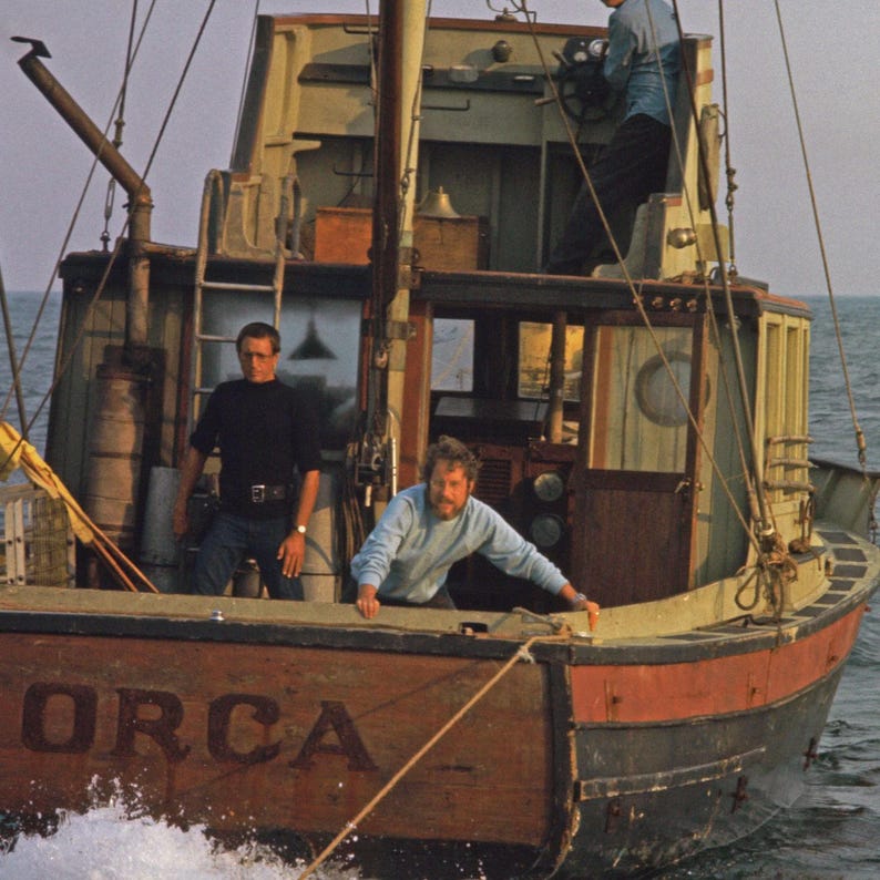 May include: A wooden fishing boat, the "Orca", sails on the ocean. The boat has a red and black hull, with the name "ORCA" painted on the stern. Three men are visible on the boat, one at the helm, and two others on deck.