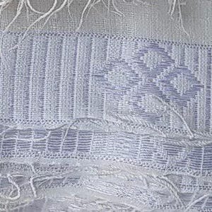 K&ouml;nnte beinhalten: Nahaufnahme eines wei&szlig;en Stoffes mit einem gewebten Muster. Der Stoff weist ein sich wiederholendes geometrisches Design in einem helleren Wei&szlig;ton mit vertikalen Streifen und Rautenformen auf. Die Kanten haben eine Fransenstruktur.