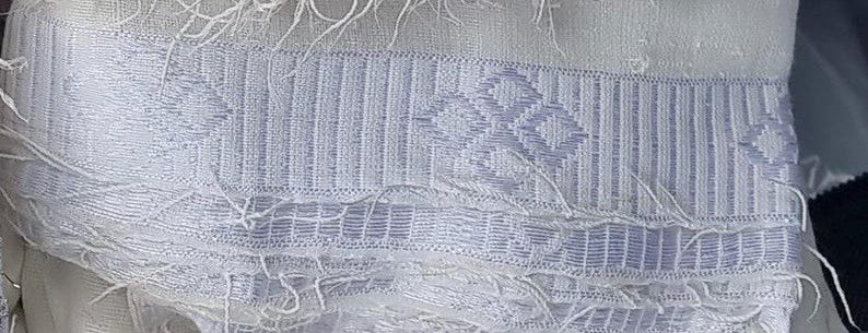 K&ouml;nnte beinhalten: Nahaufnahme eines wei&szlig;en Stoffes mit einem gewebten Muster. Der Stoff weist ein sich wiederholendes geometrisches Design in einem helleren Wei&szlig;ton mit vertikalen Streifen und Rautenformen auf. Die Kanten haben eine Fransenstruktur.