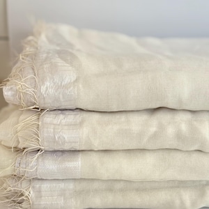 K&ouml;nnte beinhalten: Ein Stapel von vier cremefarbenen, leichten Schals. Jeder Schal hat einen Fransenrand und einen dezent schimmernden Streifen. Die Schals sind ordentlich gefaltet und zeigen ihre weiche Textur und neutrale Farbe.
