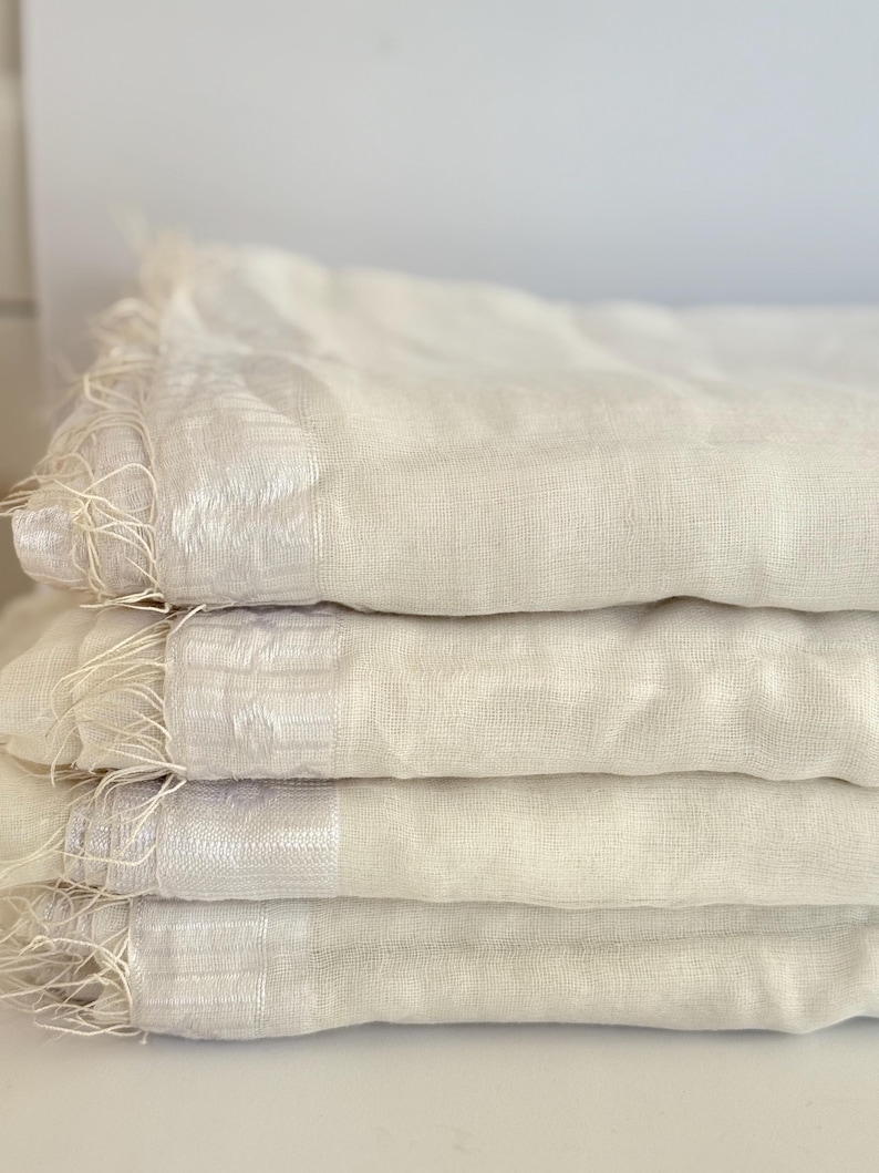 K&ouml;nnte beinhalten: Ein Stapel von vier cremefarbenen, leichten Schals. Jeder Schal hat einen Fransenrand und einen dezent schimmernden Streifen. Die Schals sind ordentlich gefaltet und zeigen ihre weiche Textur und neutrale Farbe.