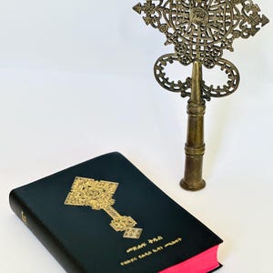 May include: A black book with gold embossed cross design and red-edged pages, next to a bronze metal processional cross. The book has text in a foreign language. The cross is ornate with a detailed openwork design.
