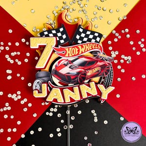 Könnte beinhalten: Eine Hot Wheels-Themen-Kuchendekoration mit der Zahl 7, einem roten Rennwagen und dem Namen "JANNY". Die Dekoration enthält karierte Flaggen, Flammen und das Hot Wheels-Logo. Der Hintergrund ist rot, gelb und schwarz.