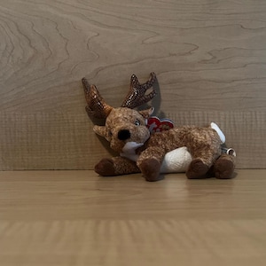 Puede incluir: Un juguete de peluche de reno marrón y blanco con una etiqueta roja y blanca que dice "Ty". El reno está acostado de lado con la cabeza girada.