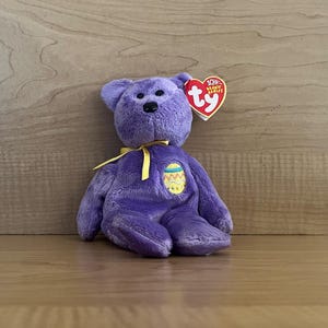 May include: Purple Ty Beanie Baby with a yellow ribbon and an embroidered Easter egg on its chest. The tag reads "Ty 10% Beanie Babies".