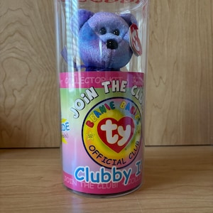 May include: A purple plush bear toy inside a clear plastic container. The bear has a black nose and eyes, and a red tag. The container has the words "Join the Club!" and "Clubby" printed on it.
