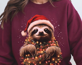 May include: Burgundy sweatshirt featuring a smiling sloth wearing a Santa hat and wrapped in Christmas lights. The sloth has brown fur and a friendly expression. Snowflakes are scattered around the sloth.