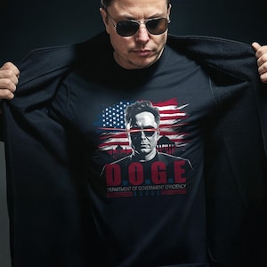 May include: A black t-shirt with a graphic of Elon Musk with a red, white, and blue American flag behind him. The text "D.O.G.E. DEPARTMENT OF GOVERNMENT EFFICIENCY" is printed below the graphic.