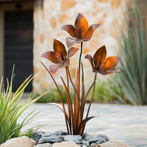 May include: A metal sculpture of three orange lilies with brown stems and leaves. The sculpture is set on a bed of gray rocks.