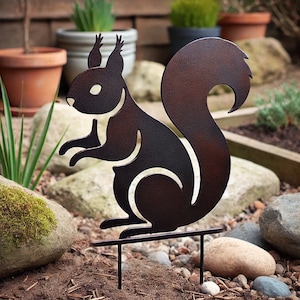 Puede incluir: Una silueta de metal de una ardilla con un acabado marrón oxidado. La ardilla está de pie sobre una estaca de metal, que se inserta en el suelo. La ardilla está mirando hacia la derecha y tiene la cola enroscada alrededor de su cuerpo.