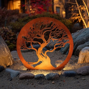 Puede incluir: Una escultura circular de metal de color naranja óxido que representa un árbol, iluminada desde atrás. Las ramas y el tronco del árbol están recortados del metal, creando una silueta. La escultura está colocada en un jardín con rocas y pequeñas luces.