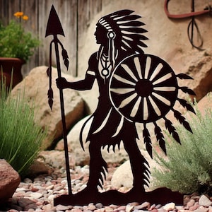 Puede incluir: Una silueta negra de metal de un guerrero nativo americano sosteniendo una lanza y un escudo. El guerrero lleva un tocado con plumas. La silueta se coloca sobre un fondo de rocas y plantas.
