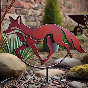 May include: A rusty red metal sculpture of a fox running, mounted on a metal stake. The sculpture is in a garden setting with rocks and green plants.