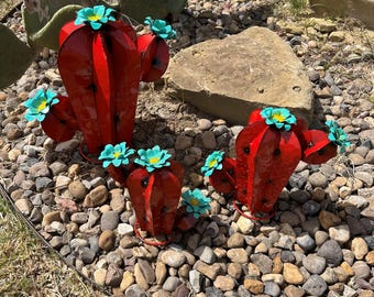 May include: Three red metal cactus sculptures with blue flowers. The cactus sculptures are on a bed of small rocks.