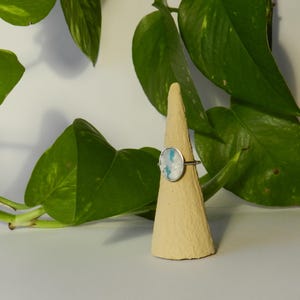 Op de afbeelding: Een zilveren ring met een ovale, lichtblauwe en witte steen wordt getoond op een beige, kegelvormige ringhouder. De ring staat tegen een achtergrond van groene bladeren en een wit oppervlak.