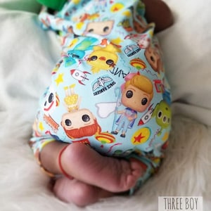 Könnte beinhalten: Ein Baby trägt einen blauen und weißen Strampler mit einem Toy Story-Charakterdruck. Der Strampler zeigt verschiedene Charaktere aus dem Film, darunter Woody, Buzz Lightyear und Jessie. Die Füße des Babys sind am unteren Rand des Bildes zu sehen.