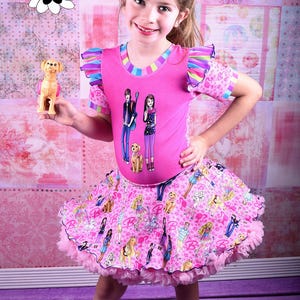 May include: A young girl wearing a pink and rainbow striped short-sleeved shirt with a Barbie graphic print. She is also wearing a pink tulle skirt with a Barbie print. The skirt has a ruffled trim. The girl is holding a small toy dog.