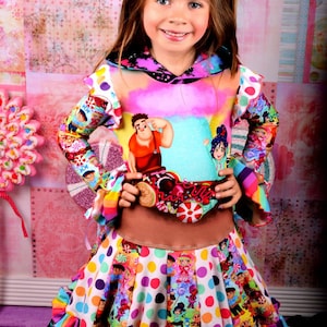 May include: A young girl wearing a colorful dress with a cartoon character print. The dress has a pink and white polka dot skirt with a rainbow ruffle trim. The top of the dress features a cartoon character from the movie Wreck-It Ralph. The girl is wearing a pink bow in her hair.