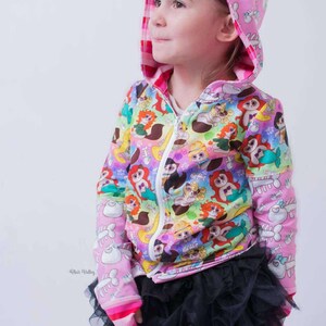 Könnte beinhalten: Eine rosa Kapuzenjacke mit Reißverschluss und buntem Cartoon-Print, kombiniert mit einem schwarzen Tutu-Rock. Die Jacke hat einen weißen Reißverschluss und rosa Bündchen. Die Kapuze hat ein sich wiederholendes Muster aus weißen Cartoonfiguren auf rosa Hintergrund.
