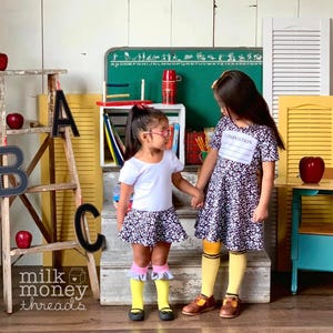 May include: Two young girls holding hands, wearing school-themed outfits. One girl wears a white top and floral skirt with pink and yellow socks. The other wears a dress with the word "COMPOSITION" and yellow socks. Apples and a chalkboard are in the background.