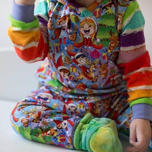 May include: A child wearing a blue, green, red, and yellow striped long-sleeved pajamas with a Christmas elf print. The pajamas feature a variety of elves, reindeer, and Christmas trees.