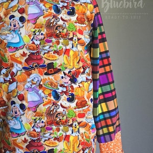 May include: A long-sleeved orange and white sweatshirt with a colorful plaid pattern on the sleeves. The front of the sweatshirt features a Thanksgiving-themed print with Disney characters like Mickey Mouse, Donald Duck, and Goofy dressed as pilgrims. The print also includes images of pumpkins, turkeys, and pies.