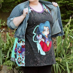 May include: A black apron featuring a cosmic design and a cartoon illustration of a villain with blue hair and a cat. The apron has a purple trim and a turquoise and black patterned side panel. The model is wearing a denim jacket and black leggings.