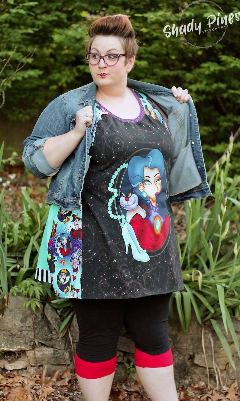 May include: A black apron featuring a cosmic design and a cartoon illustration of a villain with blue hair and a cat. The apron has a purple trim and a turquoise and black patterned side panel. The model is wearing a denim jacket and black leggings.