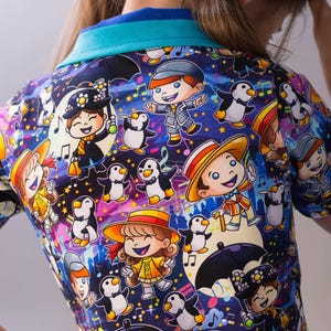 Puede incluir: Una colorida camiseta de manga corta con un estampado de dibujos animados caprichoso. El diseño presenta personajes, pingüinos, notas musicales y estrellas sobre un fondo azul oscuro. La camiseta tiene un cuello turquesa.