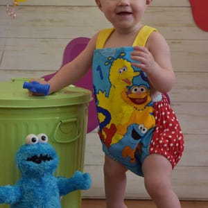 May include: A toddler girl wearing a blue and yellow Sesame Street themed romper with red polka dots. She is standing next to a green bin with a blue Sesame Street character plush toy in front of it. The girl is wearing a headband with a blue and green bow.