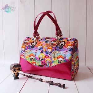 May include: A pink and patterned handbag featuring a colorful print of cartoon characters. The bag has a matching pink base and handles. A Harry Potter figurine and wand are in front of the bag. The bag has the text "Shannon Created".