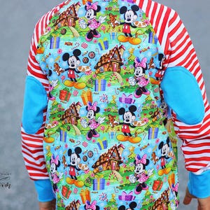 Op de afbeelding: Een shirt met lange mouwen met een kleurrijk patroon van Mickey en Minnie Mouse, peperkoekhuizen en cadeaus. De mouwen hebben rood-witte strepen, met blauwe elleboogstukken en manchetten.