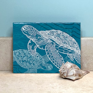 May include: A teal square art print featuring two white sea turtles with intricate patterns. The artwork is displayed next to a seashell. The background is a light blue.