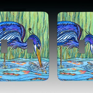 May include: A pair of light switch covers featuring a blue heron with a long beak, standing in a pond with green reeds and orange fish. The heron is depicted with detailed patterns and colors.